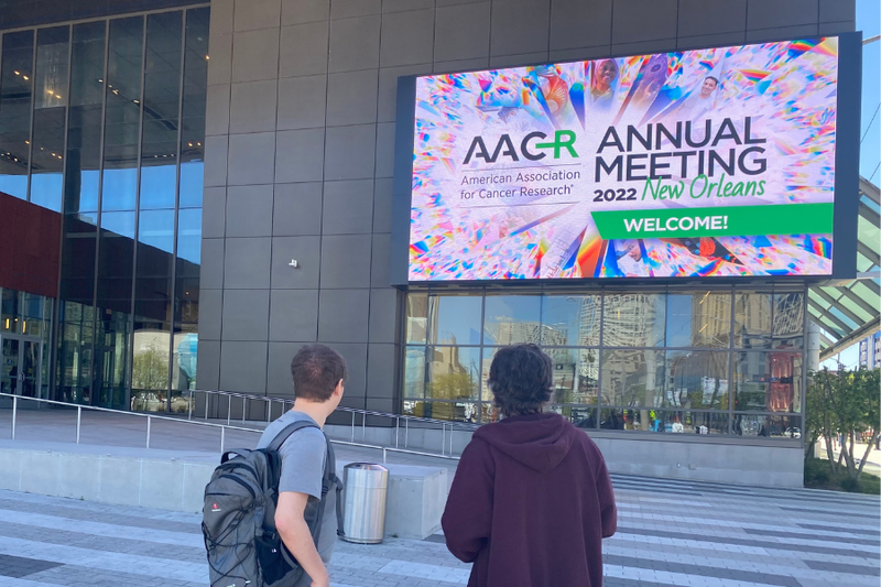 Steve and Ian stood in foreground facing the AACR Sign which is big up on a screen on the wall of a conference centre